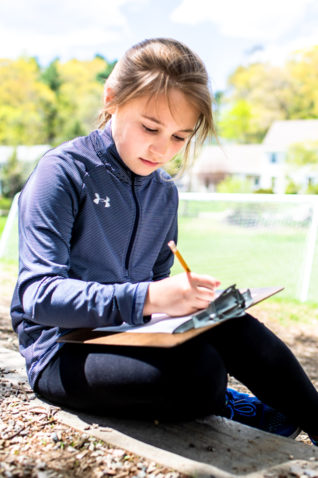 A student sitting outside writing on a clipboard.
