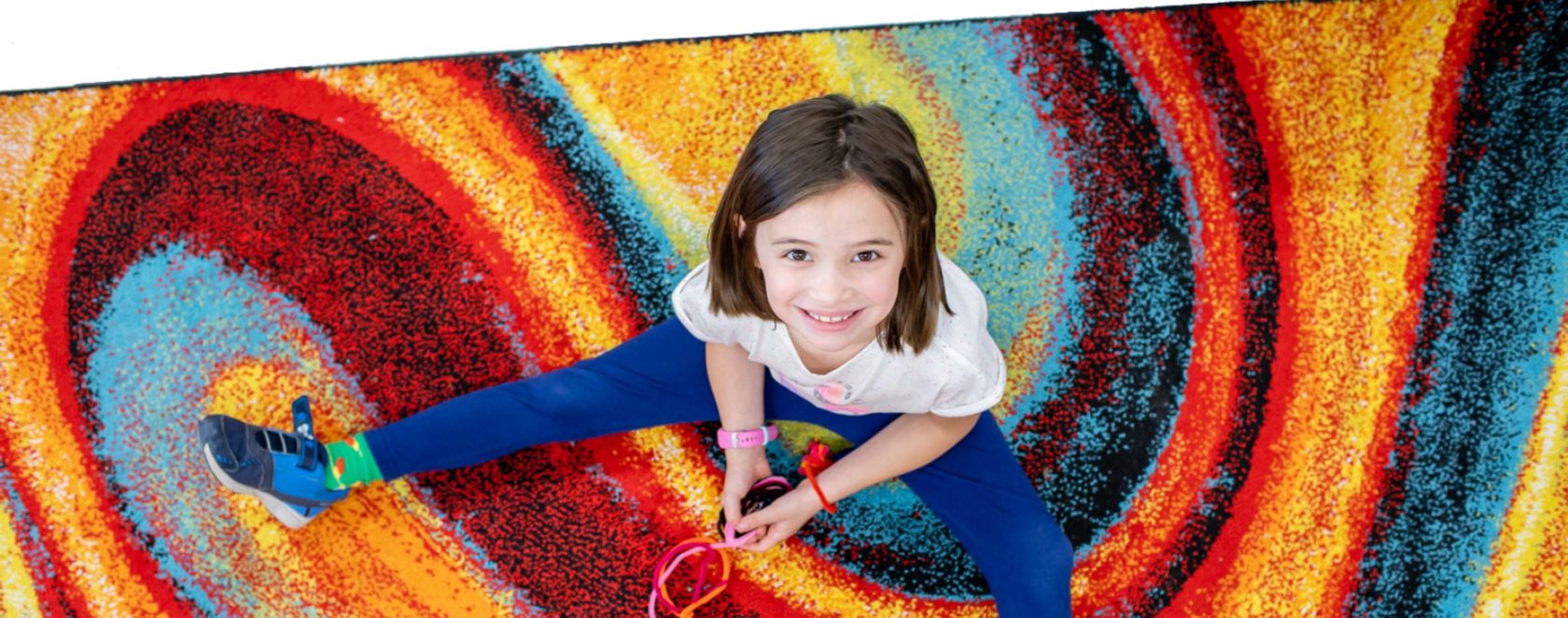 A student sitting on a colorful rug smiling and looking up at the camera.
